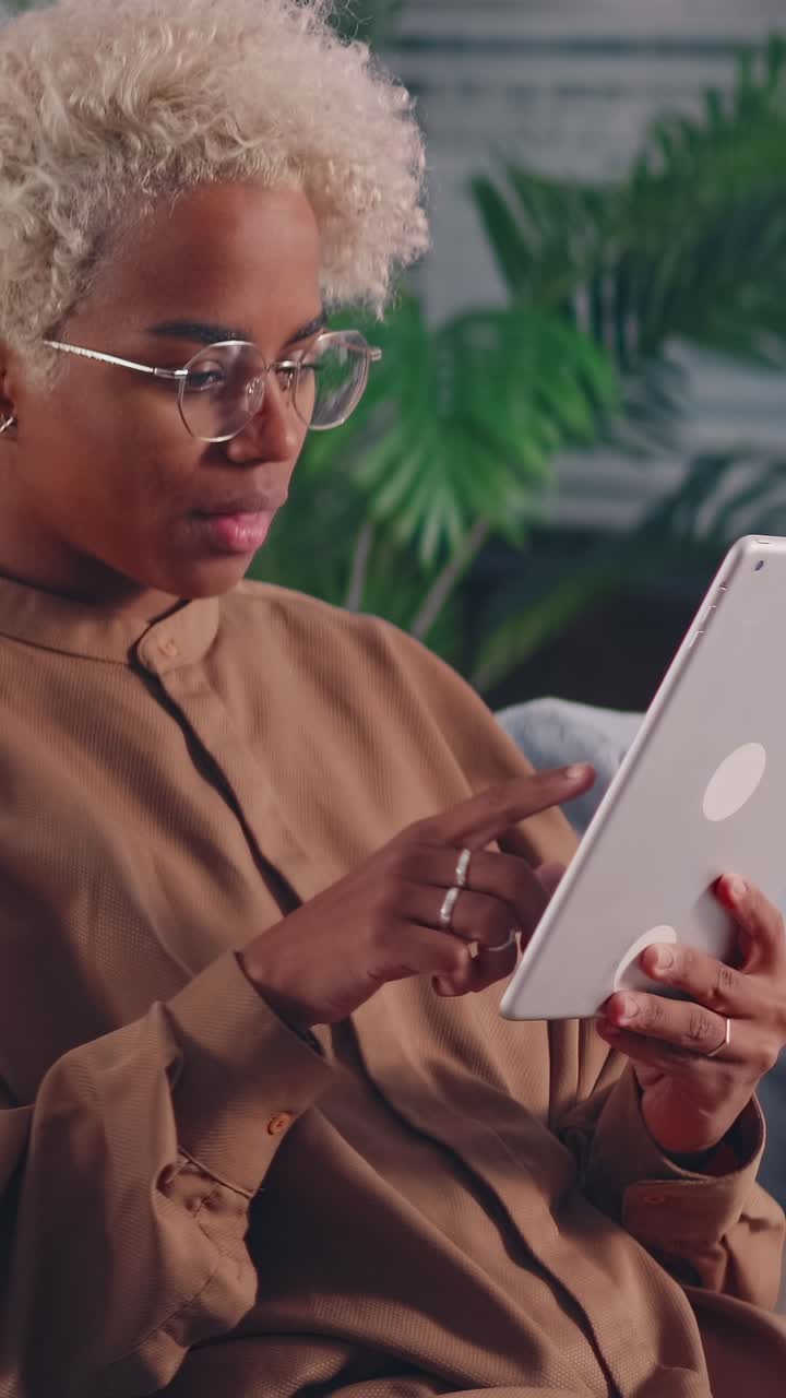 Young african american woman touches screen of electronic tablet sits on sofa