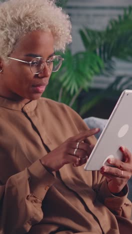 young african american woman touches screen of electronic tablet sits on sofa