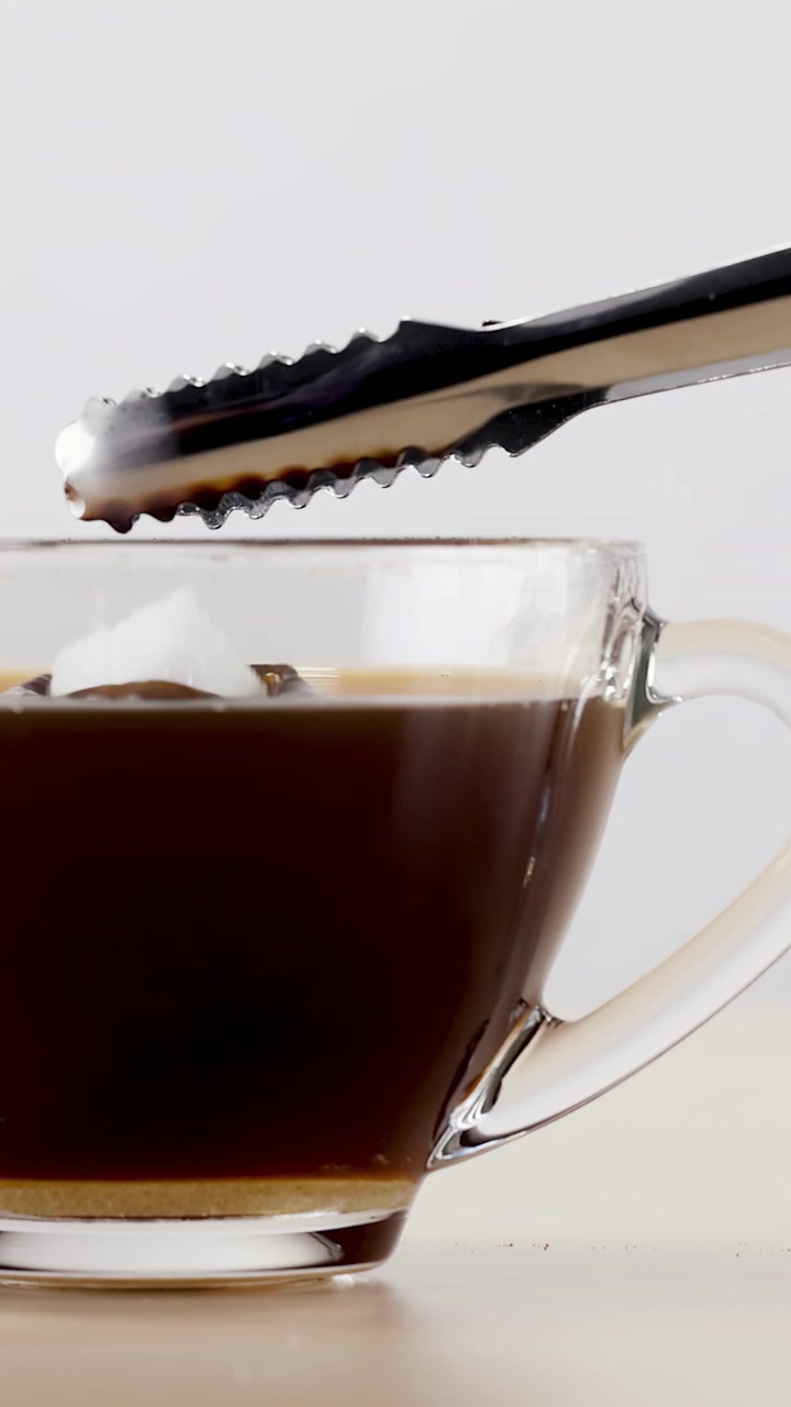 Primer plano de pinzas metálicas dejando caer un terrón de azúcar en una taza de cristal de café negro.
