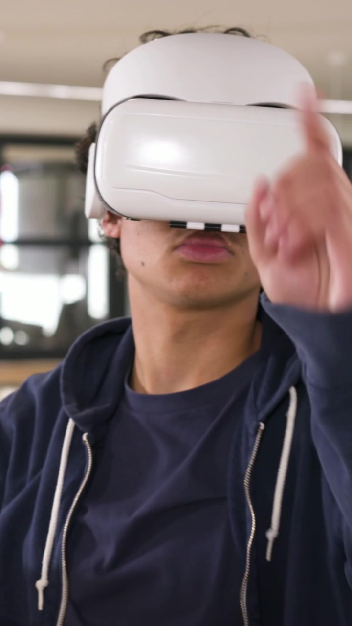 Video verticale: Uomo che indossa un visore VR bianco, con felpa blu navy, che testa i controlli VR in un ufficio open space.