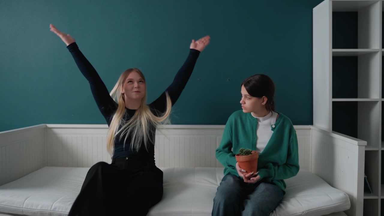 Two women sitting on daybed in new apartment discussing unpacking, plant placement, teal wall, empty shelves, mattress on frame, calm reflective mood, gesturing, quiet laughter, planning decor