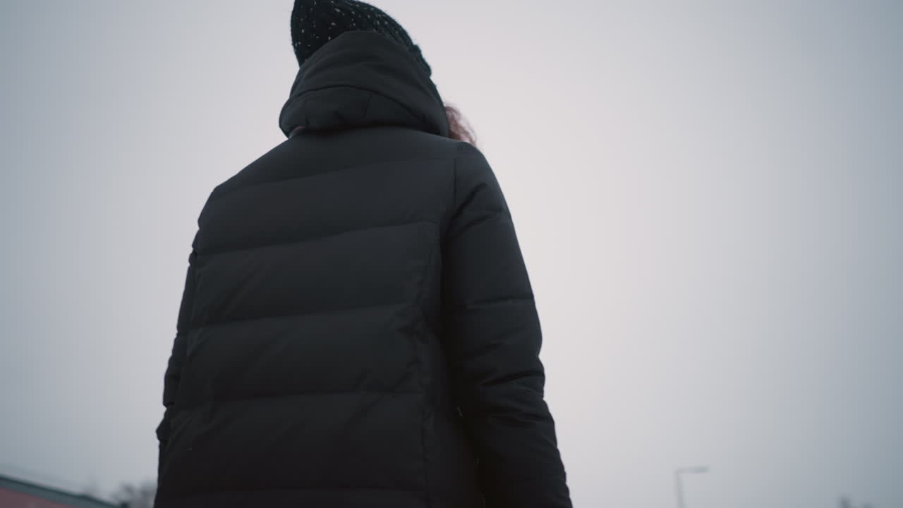 Lady dressed in black winter coat and knit hat standing outdoors during cold cloudy day, close up of shoulder and head with background of snowy trees and sky creating seasonal atmosphere