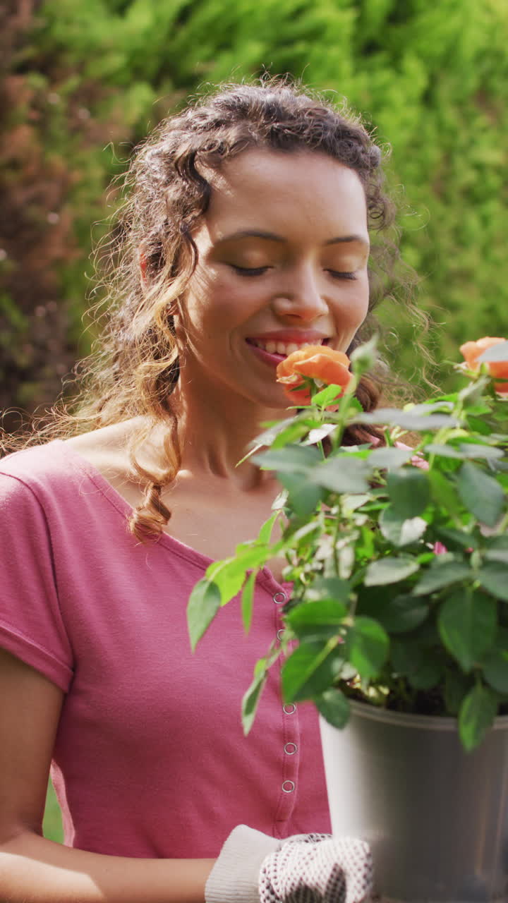 video vertical de una mujer biracial feliz sosteniendo flores sonriendo en el jardín