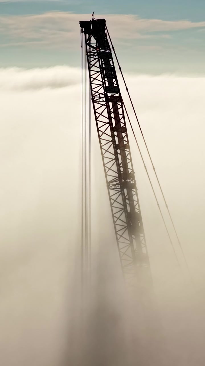 Inquadratura verticale surreale di una grande torre di gru industriale a traliccio verde e arancione che si innalza alta sopra una fitta coltre di nebbia bassa e nuvole in un cielo azzurro limpido in una mattina nebbiosa.