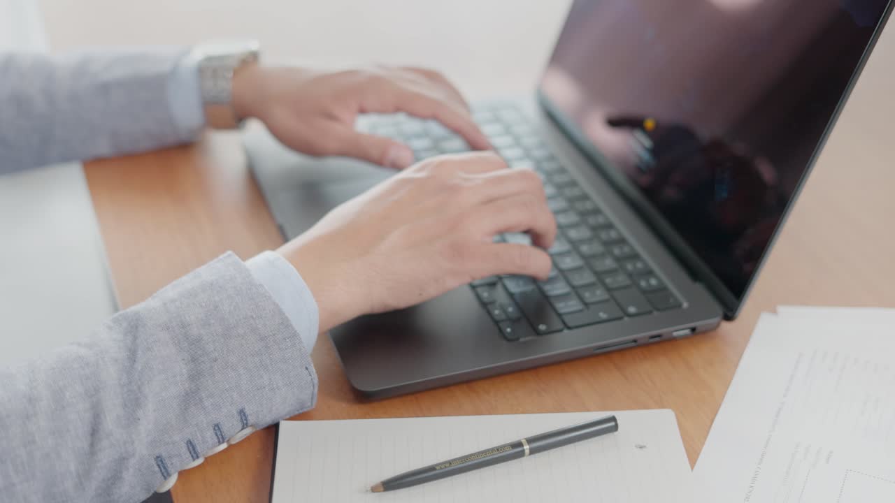 Semi orbit view of hands typing on a laptop in office setting with papers and pen on desk
