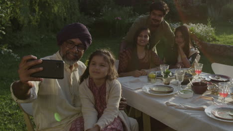 a multi-generational family takes a selfie during an outdoor meal, including a tender moment with a child's kiss