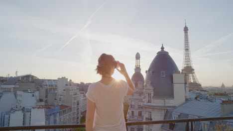 Mujer-Que-Usa-Auriculares-De-Realidad-Virtual-Y-Disfruta-Explorando-La-Experiencia-Del-Ciberespacio-En-Línea-En-El-Balcón-De-La-Hermosa-Torre-Eiffel-Al-Atardecer-De-París