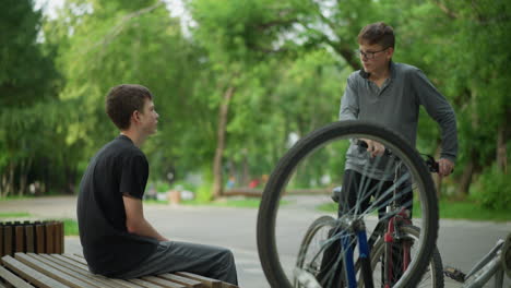 young boy seated on park bench beside an upside-down bicycle with spinning tires is approached by another boy riding bicycle, the two begin to look at each other without saying anything