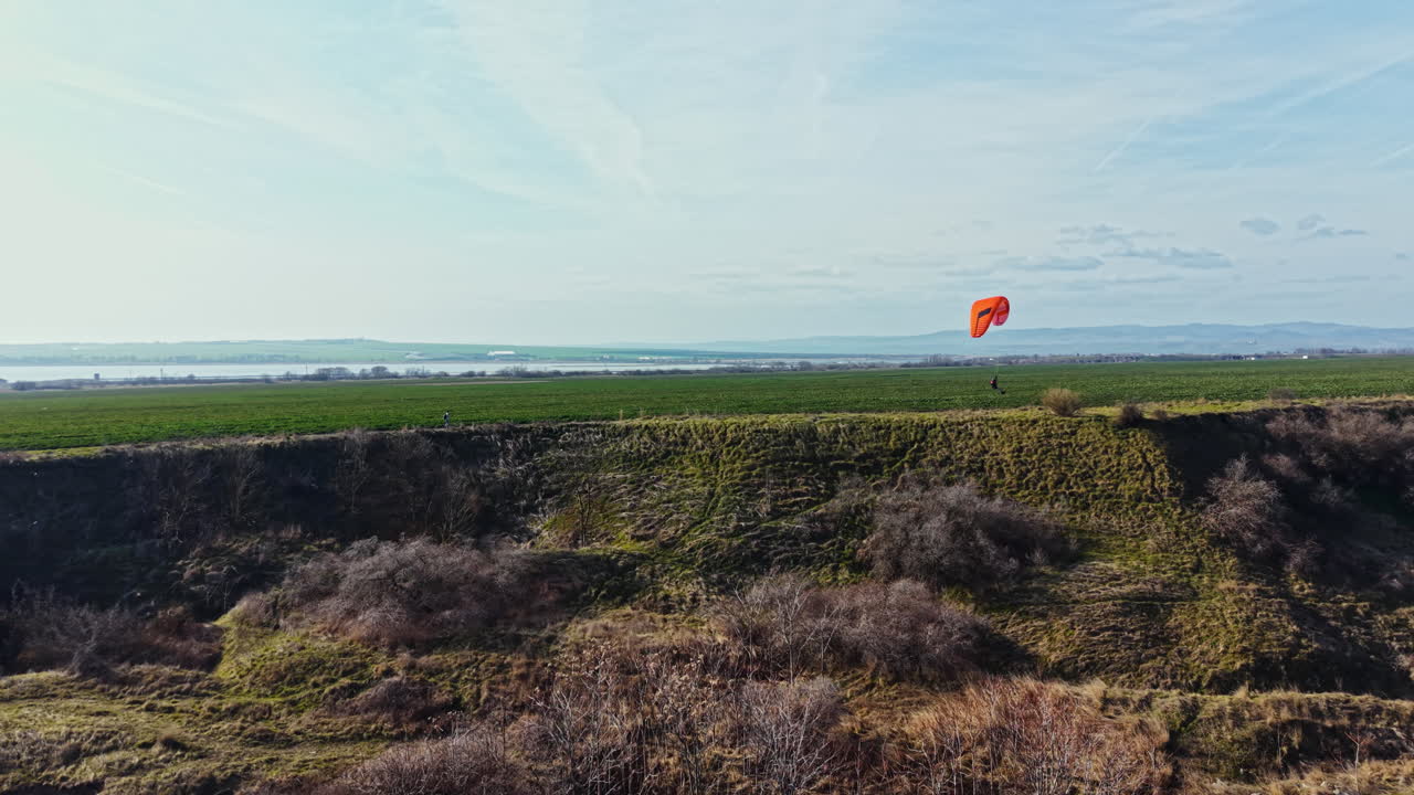 Parapente voa sobre campo verde perto de penhasco em dia ensolarado com céu azul e água ao longe