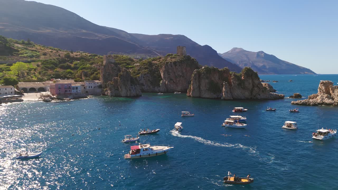 Aerial drone footage orbiting right around the historic Tonnara di Scopello, showcasing the rugged Sicilian rocky coastline and a boat on the sparkling blue ocean in Italian summer in Sicily, Italy