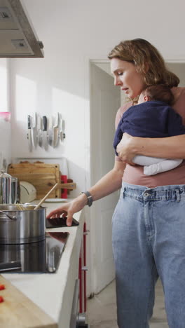 video vertical de una madre caucásica con un bebé recién nacido en un portero usando un teléfono inteligente en la cocina