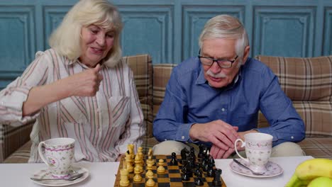 Retired-senior-couple-talking-drinking-tea,-playing-chess-in-modern-living-home-room-lounge-together