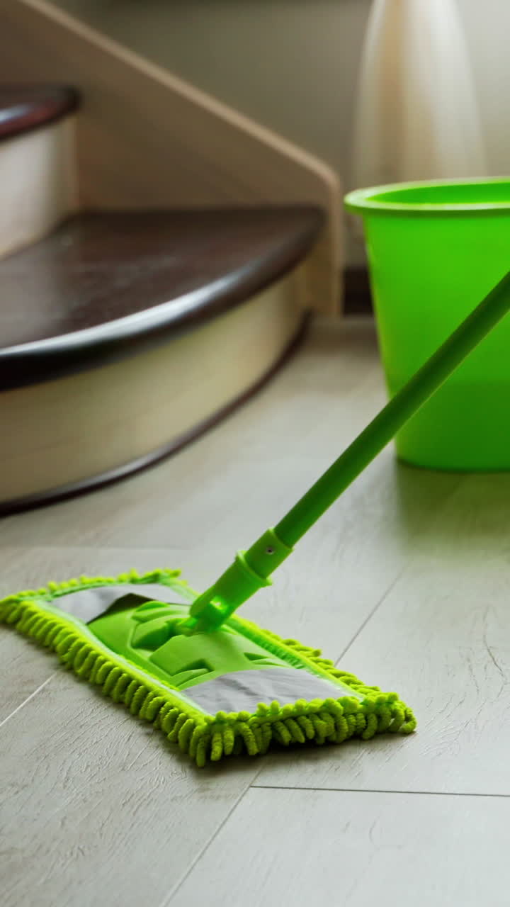 Cleaning the floor in the living-room. Microfiber mop dusting the parquet floor near the green bucket in the room. Householding chores. Vertical video