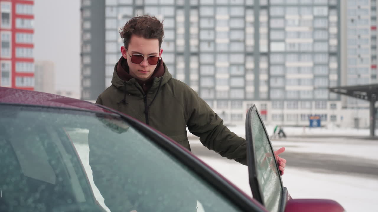 Man in winter jacket approaches parked car during cold snowy day with urban residential and office buildings in background, snow visible on vehicles and ground, indicating chilly winter weather