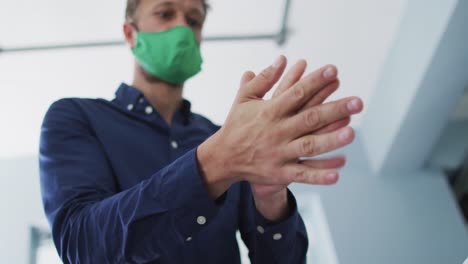 hombre caucásico con máscara facial usando desinfectante para manos en una oficina moderna