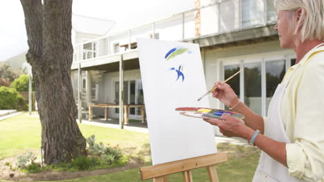 Feliz-Mujer-Mayor-Caucásica-Pintando-Cuadros-Y-Sonriendo-En-Un-Jardín-Soleado,-Cámara-Lenta