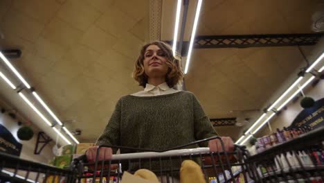 Pretty-woman-in-casual-clothes-is-walking-in-grocery-store-steering-shopping-trolley-with-bread-inside-it-and-looking-around-at-shelves-with-products.-Women-and-shops-concept.-Low-angle-footage-from-the-cart