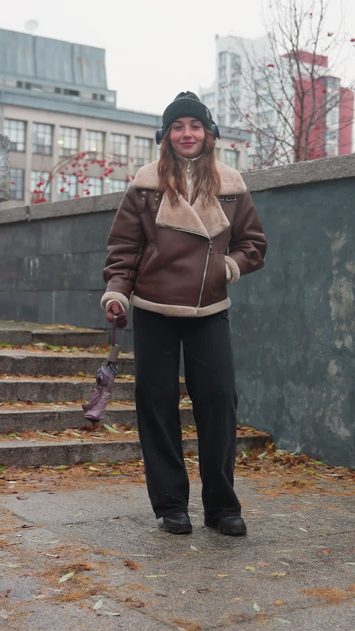 Cheerful lady wearing black knit cap, brown shearling jacket, black trousers, standing outdoors with one hand in pocket while swinging umbrella playfully on concrete path with autumn leaves scattered