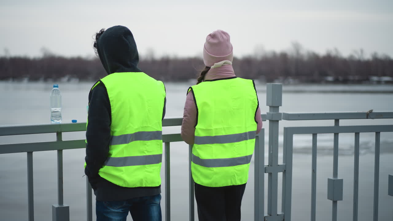Man in hoodie and woman in pink jacket wearing neon safety vests standing at railing overlooking river, man eating from container with water bottle beside, during cold overcast day