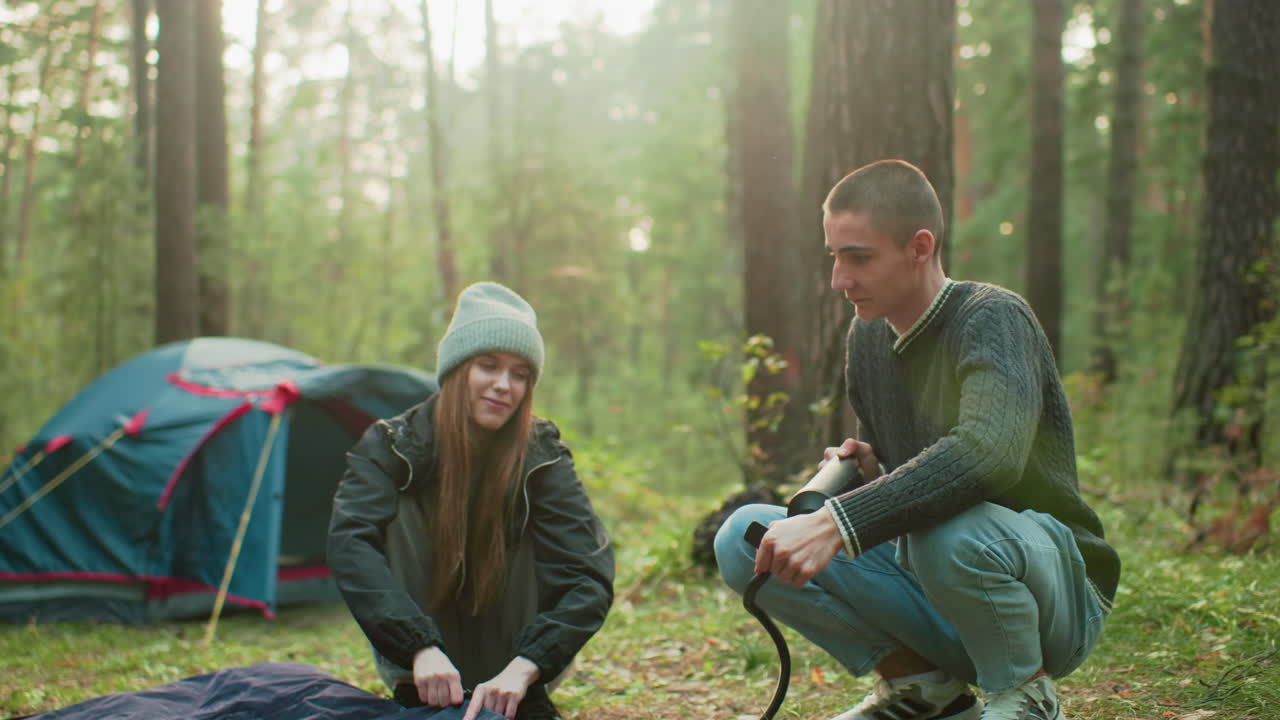 Campers enjoy outdoor moment as man pumps air into tent bag with forest backdrop and tent behind them, creating relaxed camping atmosphere filled with teamwork