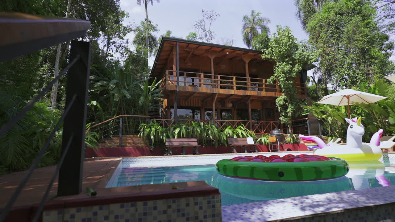 Slow reveal of jungle lodge pool scene shows unicorn and watermelon floats, wooden house, and lush greenery—captured in Selva Misionera, Misiones, Argentina with tranquil tropical atmosphere