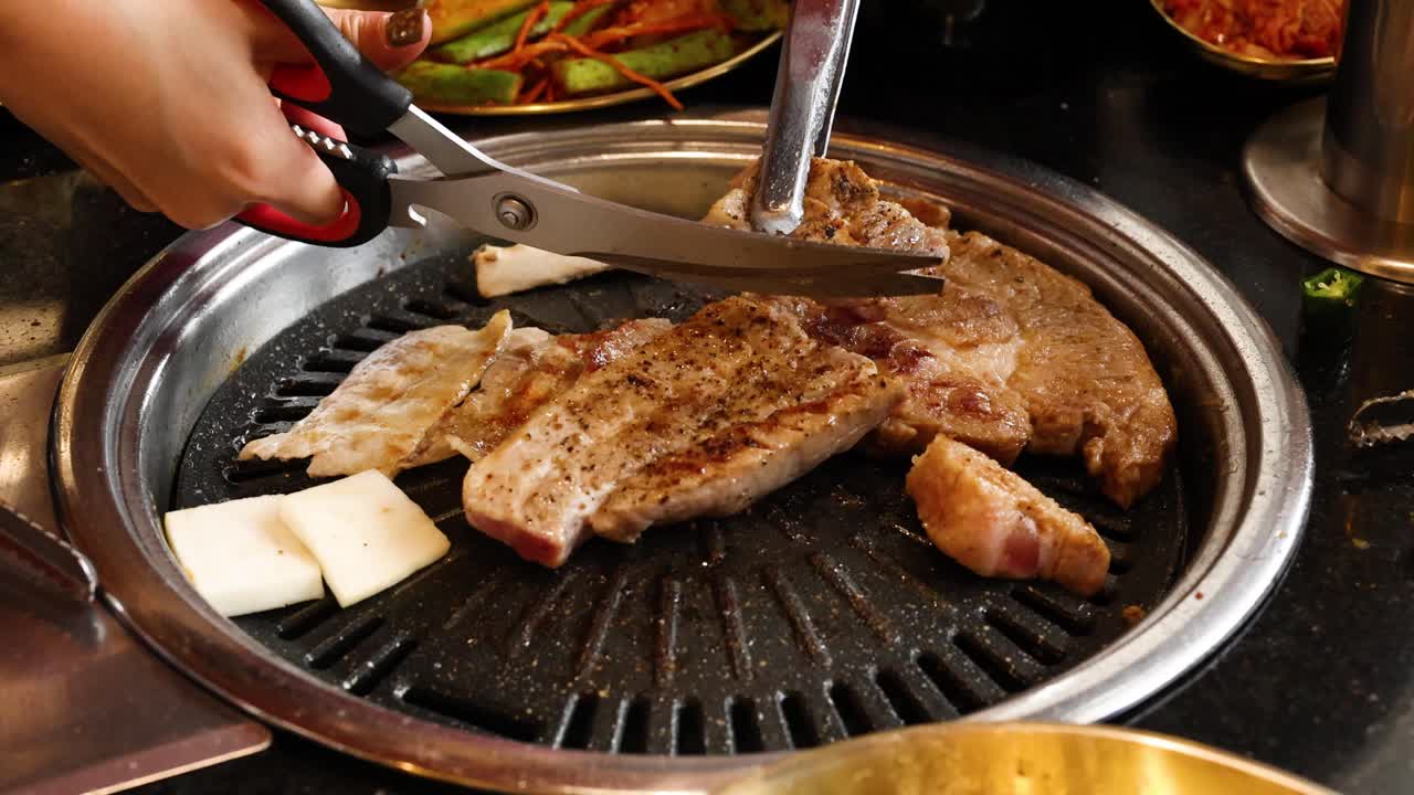 Hand cuts grilled pork belly with scissors on Korean barbecue grill under warm ambient lighting