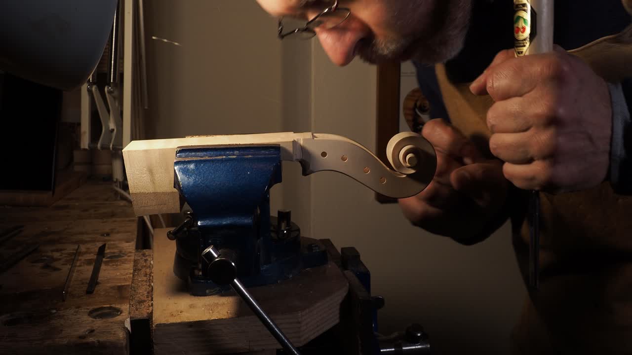 Violin maker crafts the delicate scroll and pegbox from a single piece of maple using precise hand tools, with the work secured in a vise; the image captures the focus and manual skill required