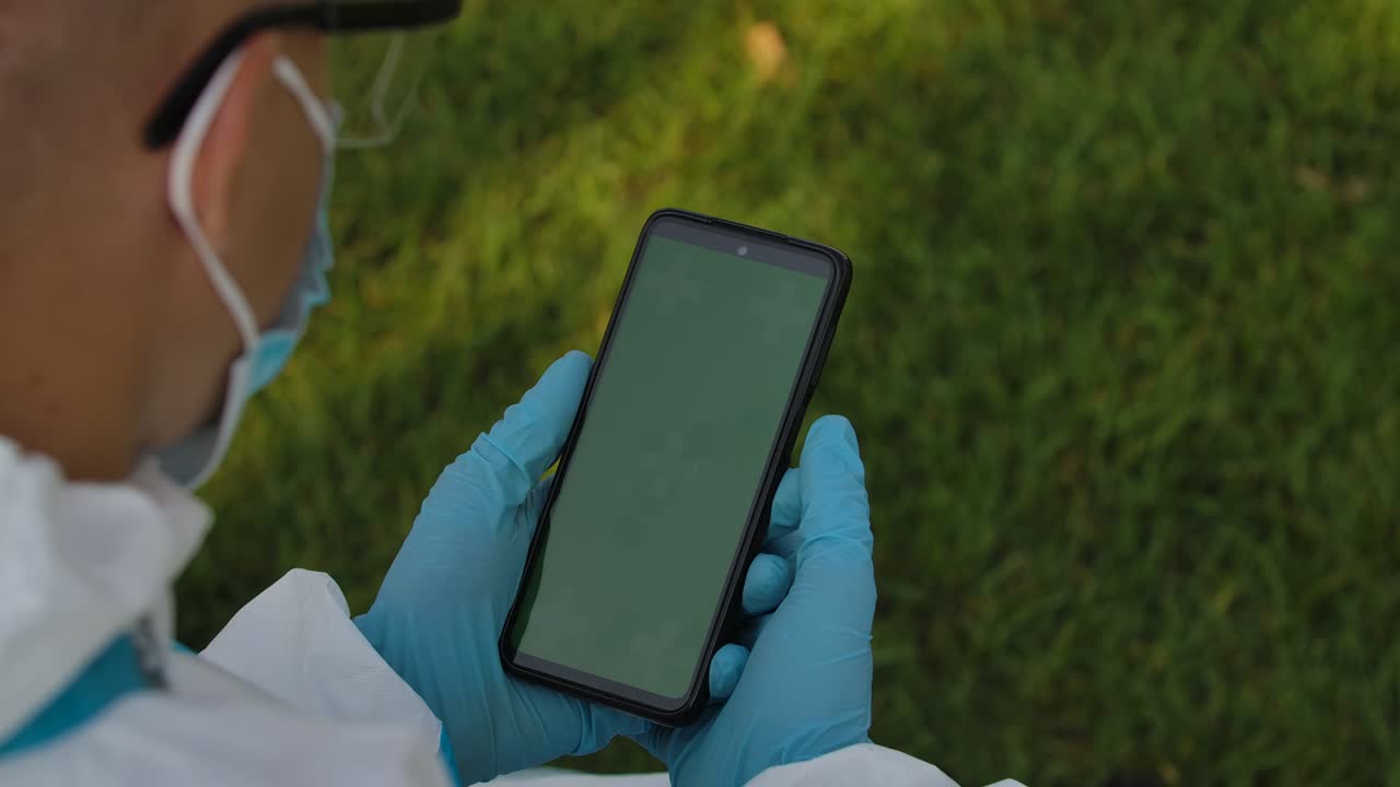 una vista detrás del hombro de un hombre con traje de protección sosteniendo un teléfono inteligente con una pantalla verde con una tecla de croma, navegando por el contenido tocando o deslizando. cerrar. movimiento lento