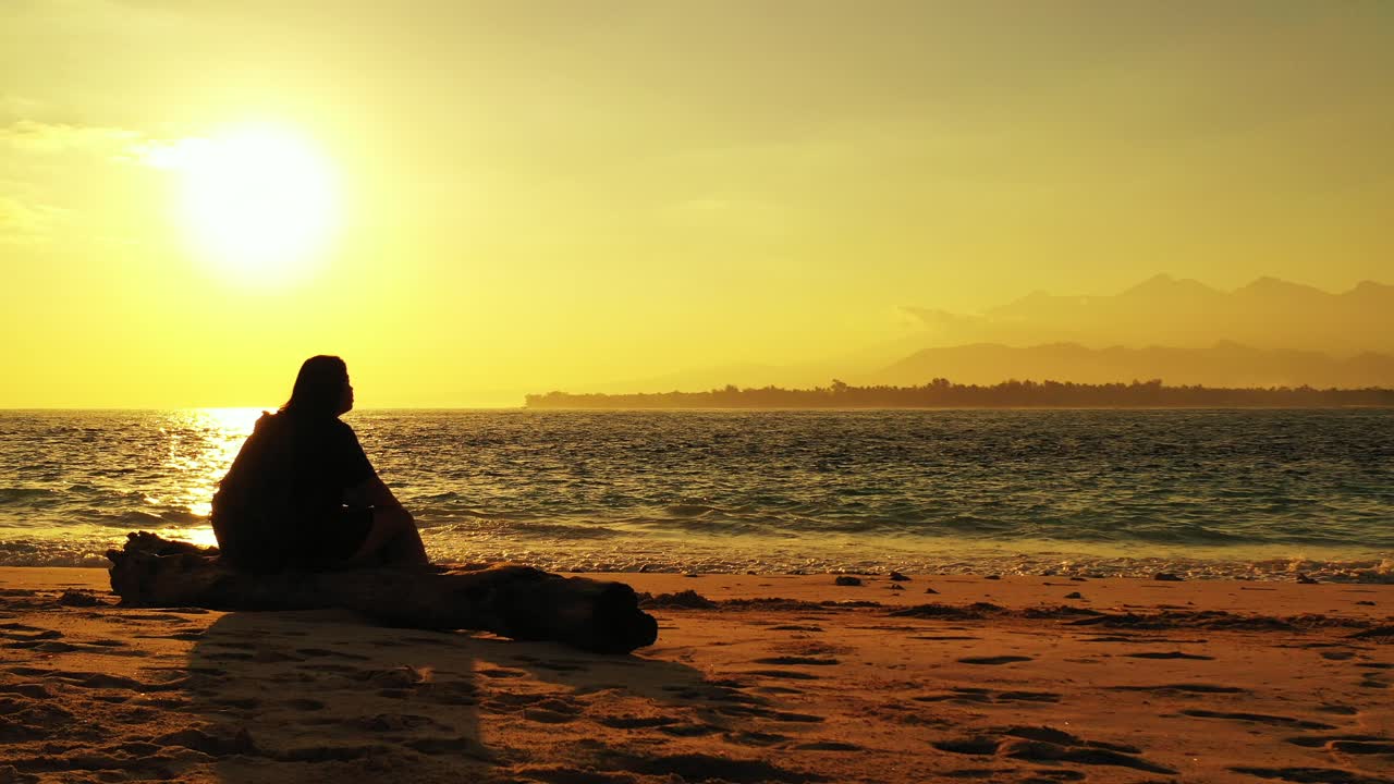 Pensive woman sitting on the old trunk on a sandy beach during the sunset. figuring out the next life step