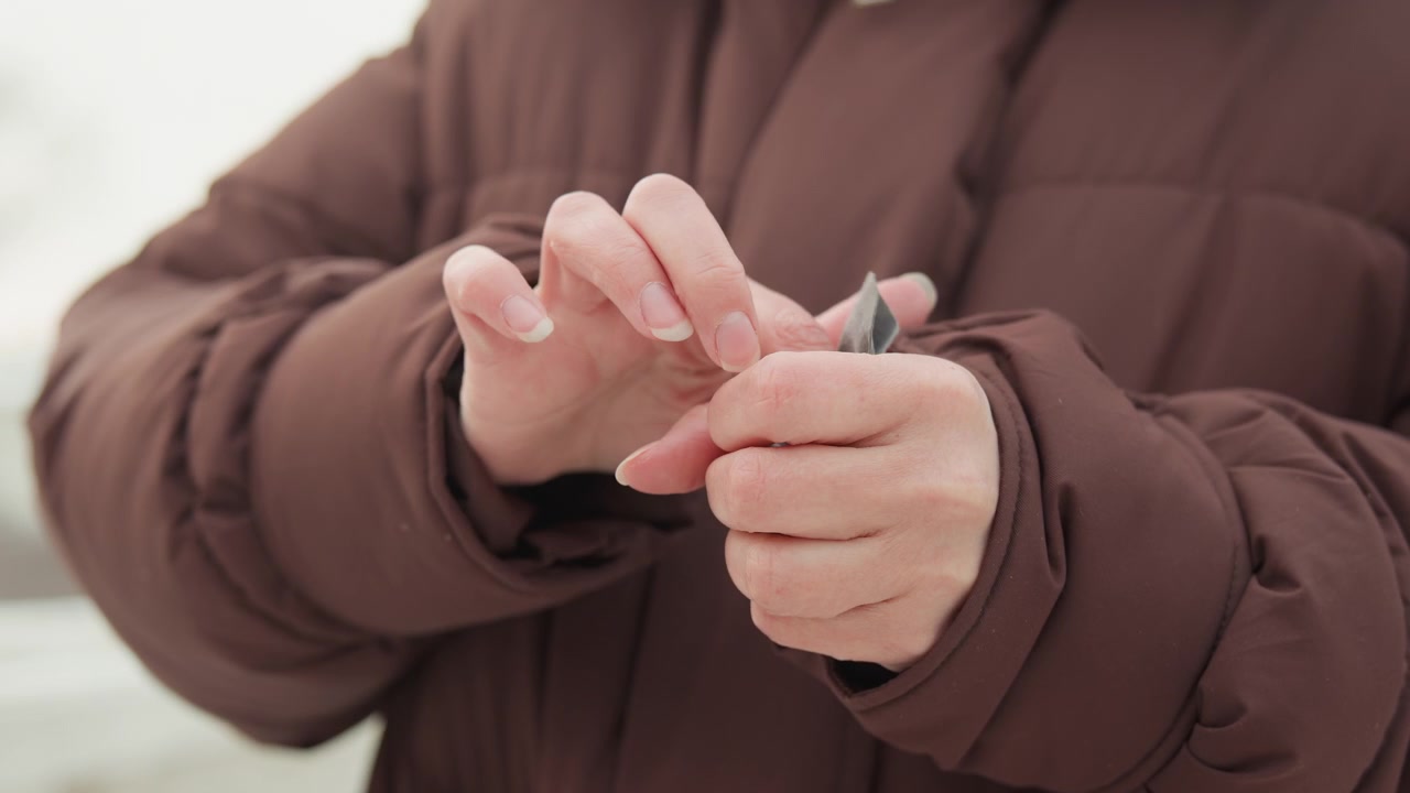 Una vista ravvicinata mostra una persona con una giacca imbottita marrone che infila un piccolo guanto di maglia grigio sulla mano destra mentre la mano sinistra liscia il polsino, con le dita che lavorano con attenzione in una luce esterna soffusa con un movimento sottile e