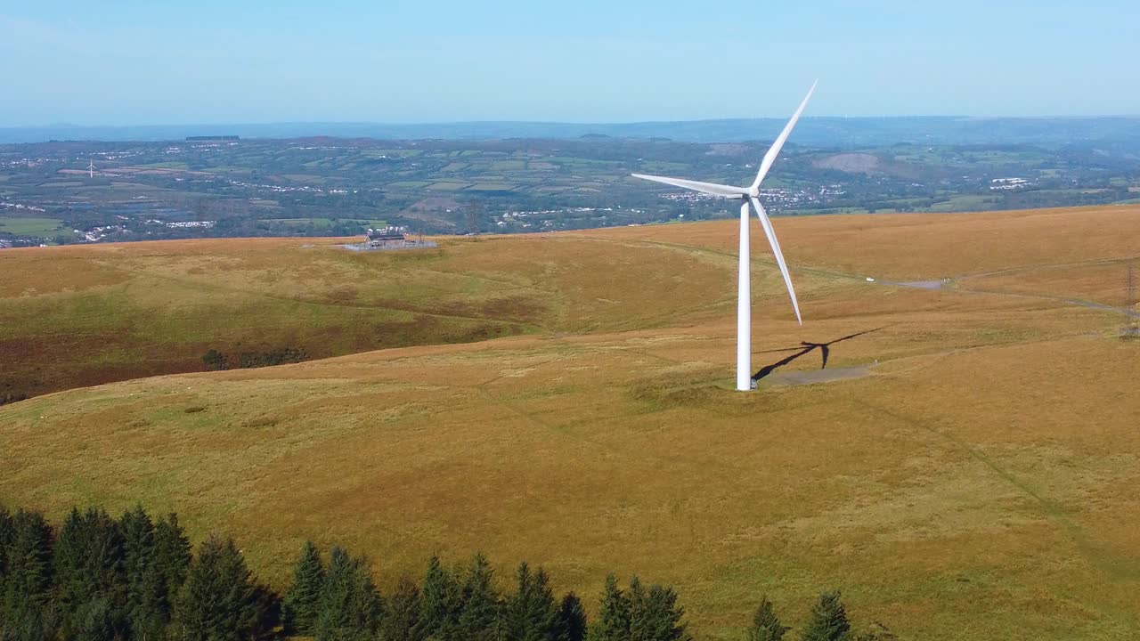 Descending Aerial Drone View of Lone Wind Turbine Spinning with Pine Trees Below and Town and Fields in Background. Eco-Friendly Renewable Energy Generation Concept Footage 4K
