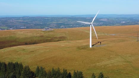 descending aerial drone view of lone wind turbine spinning with pine trees below and town and fields in background. eco-friendly renewable energy generation concept footage 4k