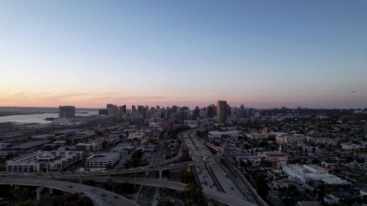 Drone recuando acima da autoestrada ao pôr do sol, revelando uma vista panorâmica do horizonte do centro de San Diego e da cidade circundante.