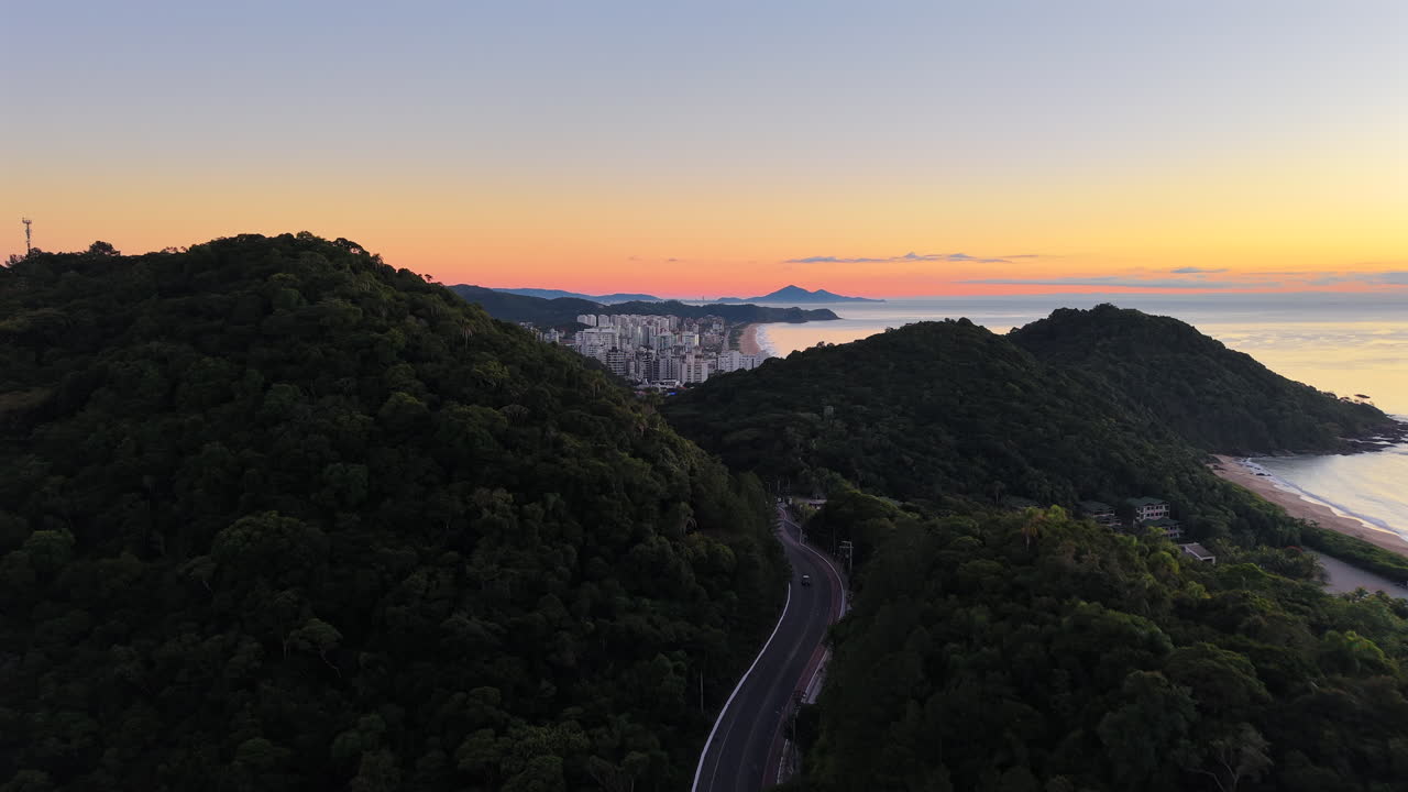 Aerial drone shot at sunrise captures lush green hills in the foreground, with the distant skyline and curved beachfront of Itajaí city visible beyond Praia do Buraco and Praia Brava, Brazil