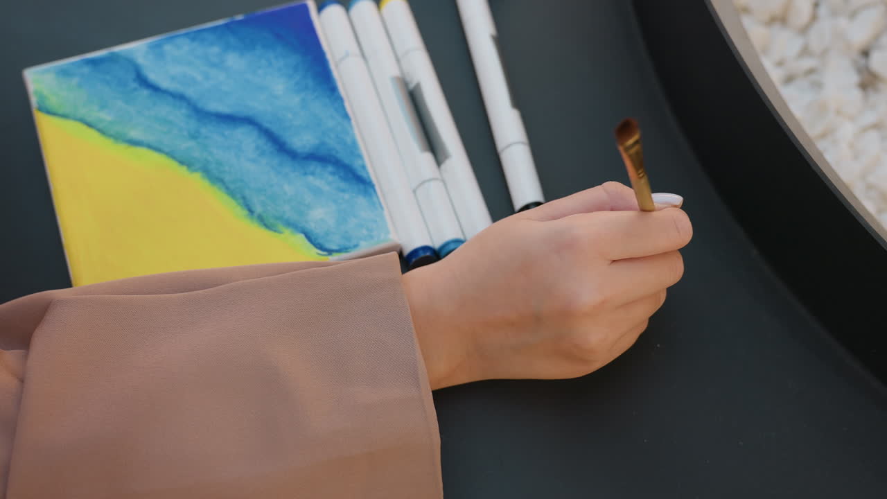 Pequeño lienzo con un paisaje marino vibrante: un artista representa el horizonte costero con colores y herramientas en capas; una pintora diseña un paisaje marítimo en un lienzo compacto empleando diversos materiales artísticos.