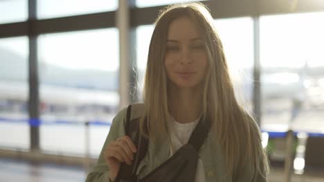 Portrait-of-smiling-young-blonde-girl-traveller-with-backpack-stands-in-airport-terminal