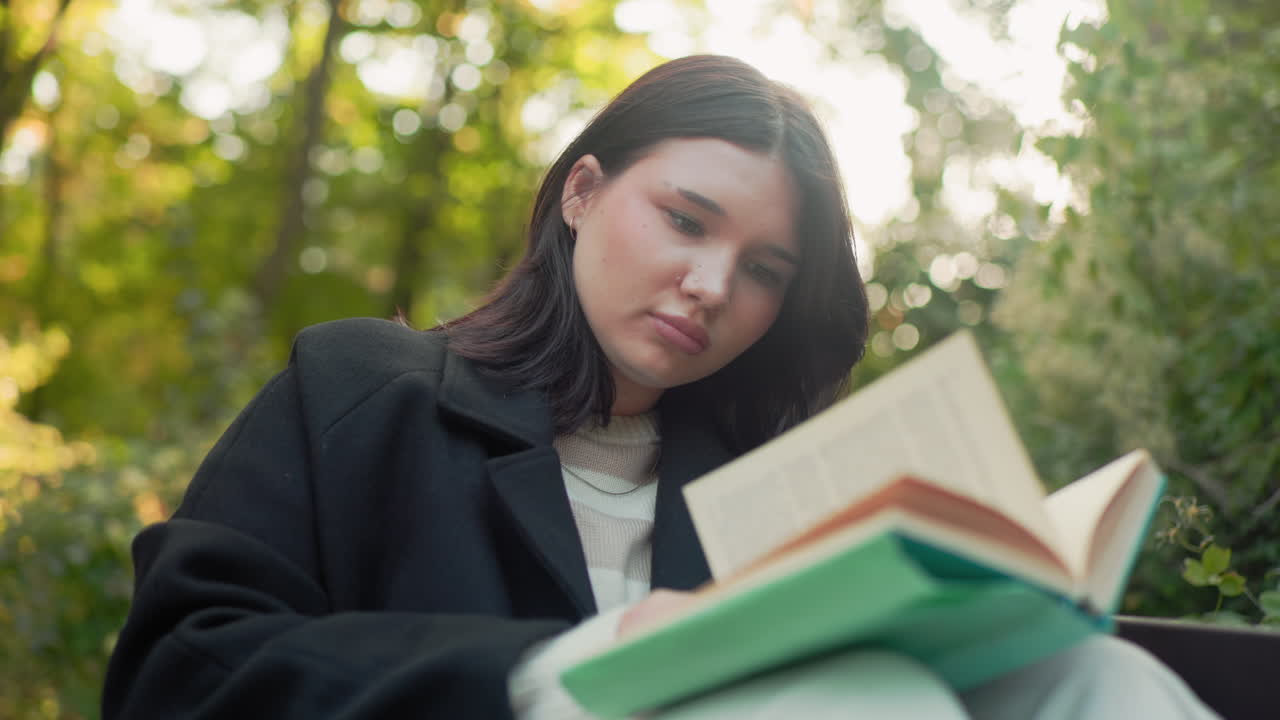 Lower view of outdoor lover seated on park bench with legs up reading book, adjusting hair between pages, paper cup on ground, black coat and white trousers, calm autumn greenery, soft sunlight bokeh
