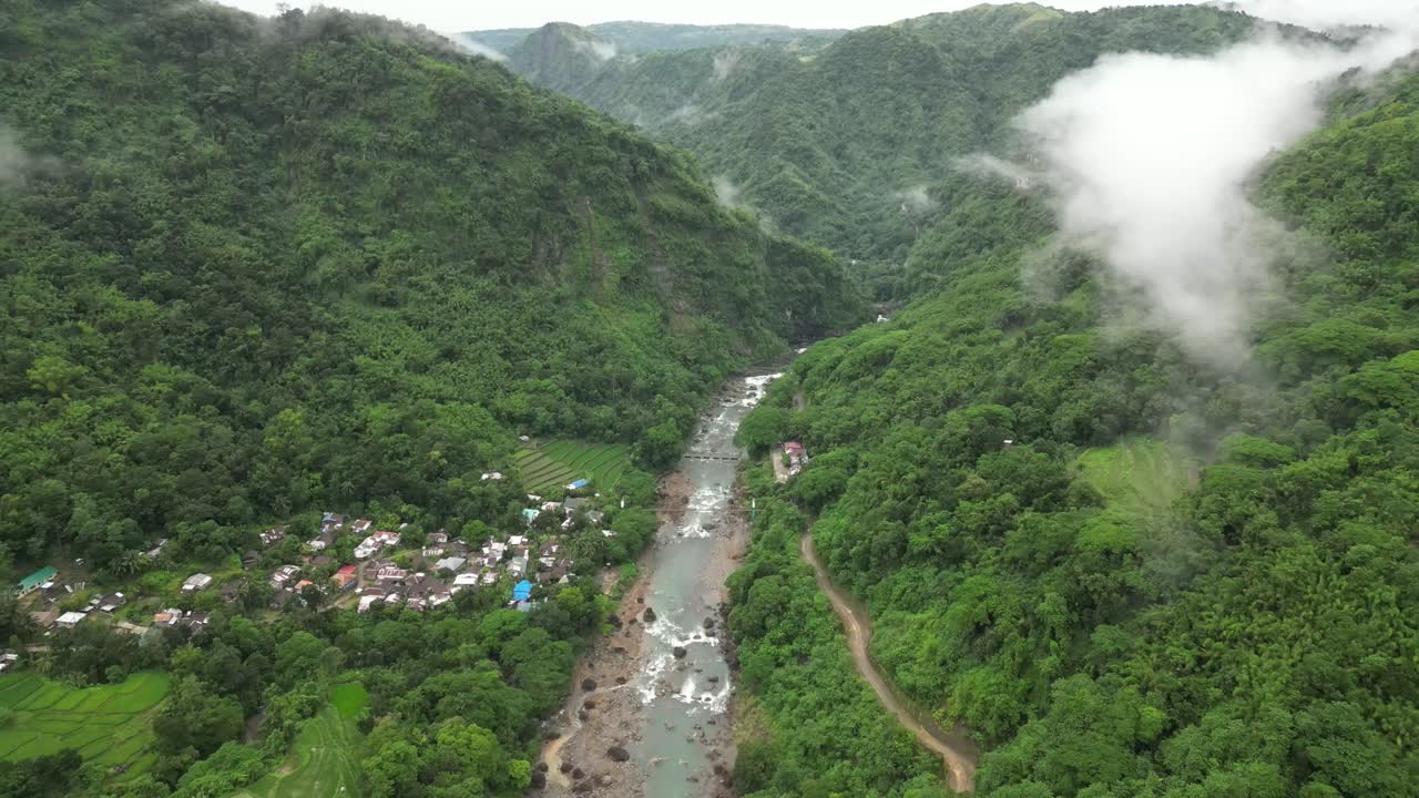 A wide aerial angle drifts to a final hold over forested mountains and a winding river. Mist threads through valleys as terraced fields and a clustered village settle into the lush terrain
