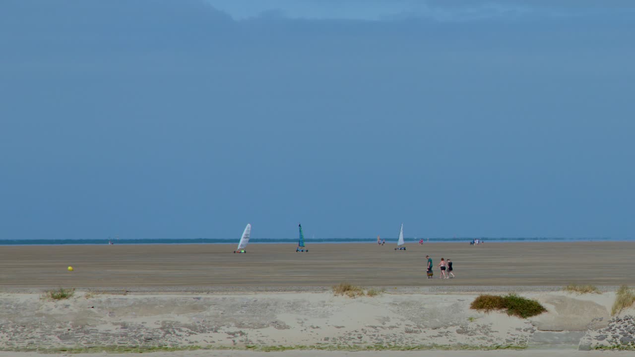 Two tourists walk a wide sandy beach as land sailers glide by under clear skies