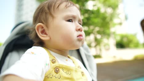 little 1 year baby girl staring at kids in the playground while she is sitting in a stroller with a serious face expression, close-up in slow motion, low angle summer daytime