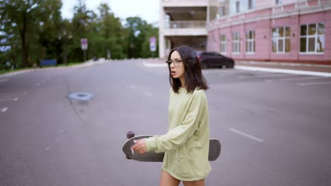 Retrato-De-Una-Morena-Con-Un-Suéter-Verde-Y-Gafas,-Que-Camina-Por-La-Carretera-Con-Una-Patineta-En-Las-Manos-Y-Fumando