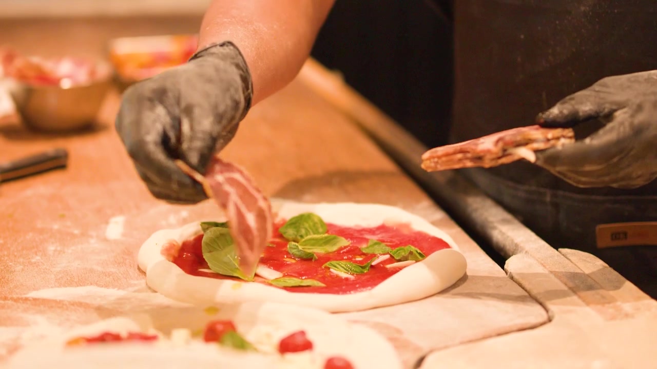 Chef con guanti neri aggiunge salumi a una pizza con salsa di pomodoro e basilico