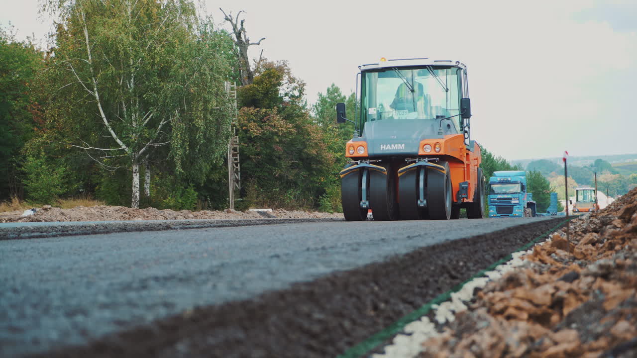 Modern asphalt roller press hot asphalt while making a new road in autumn. Road repair machine with heavy vibration rollers is working on one side of the road and other machinery going by.