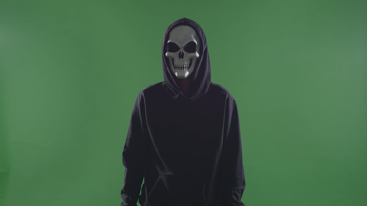 hombre aterrador en la sudadera con capucha usando máscara de halloween de pie en el fondo de la pantalla verde