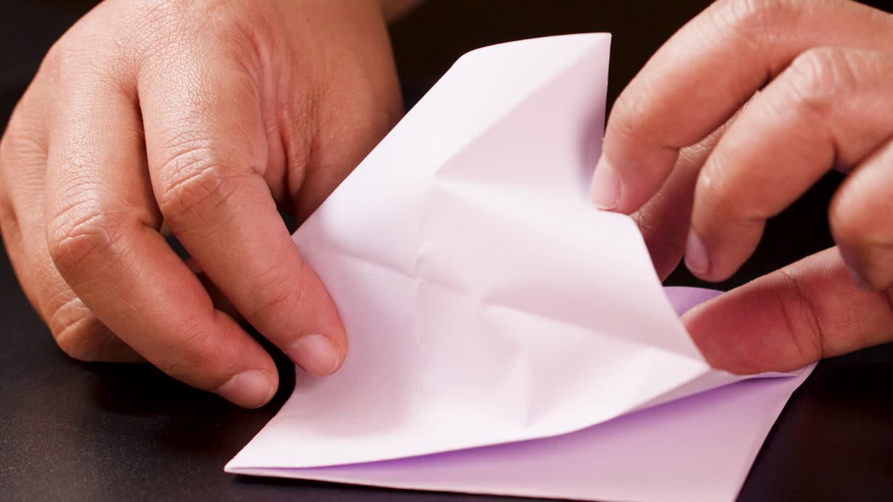 Hands carefully fold white paper into an origami boat on dark tabletop under soft lighting