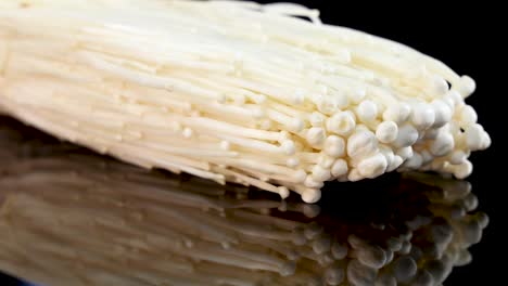 a fresh enoki mushroom cluster slowly rotates on a glossy black surface, illuminated by soft studio lighting, highlighting its delicate texture and pale color