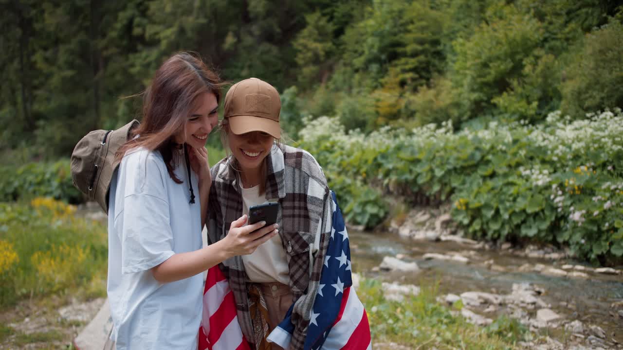 una chica morena tomó una foto de su amiga con una bandera estadounidense y ahora están mirando la foto. foto contra el fondo de un bosque verde y ríos de montaña