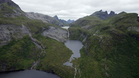 Aerial-dolly-forward-shot-of-dramatic-rocky-cliffs-and-mountains-with-a-waterfall-spilling-into-a-lake-on-an-overcast-day