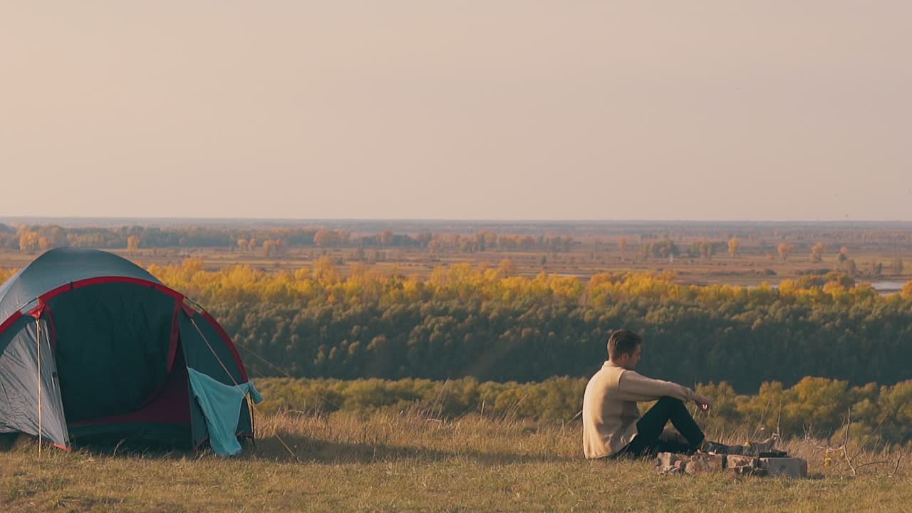 슬픈 남자가 캠프 불  ⁇ 에 앉아 가을의 숲을 바라보고 있습니다.