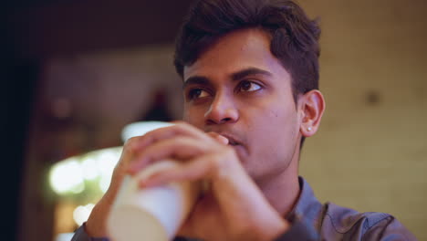 Young man with focused expression holding drink cup and adjusting straw while seated in indoor setting with colorful defocused lights, wearing buttoned shirt and wristwatch, showing calm thoughtful demeanor